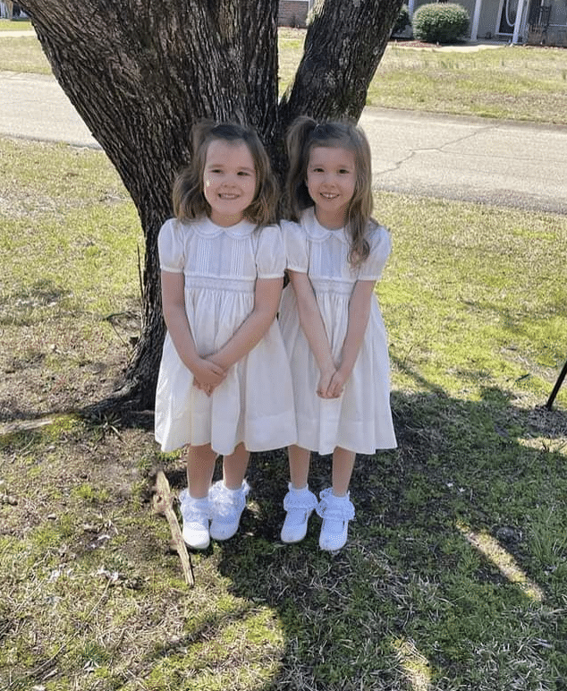 two young girls in matching off-white short sleeve dresses with white socks and dress shoes, standing in front of a tree outside on the grass