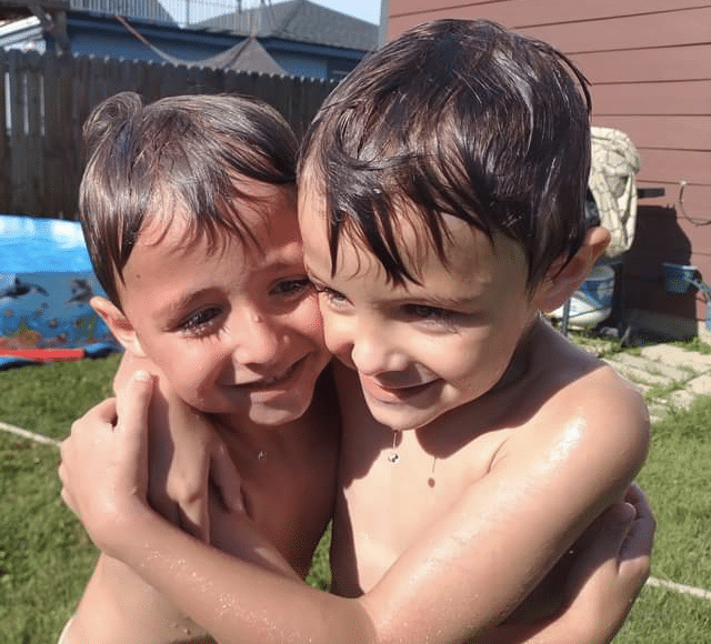 two young boys with wet, dark hair hugging each other outside with a small pool in the background