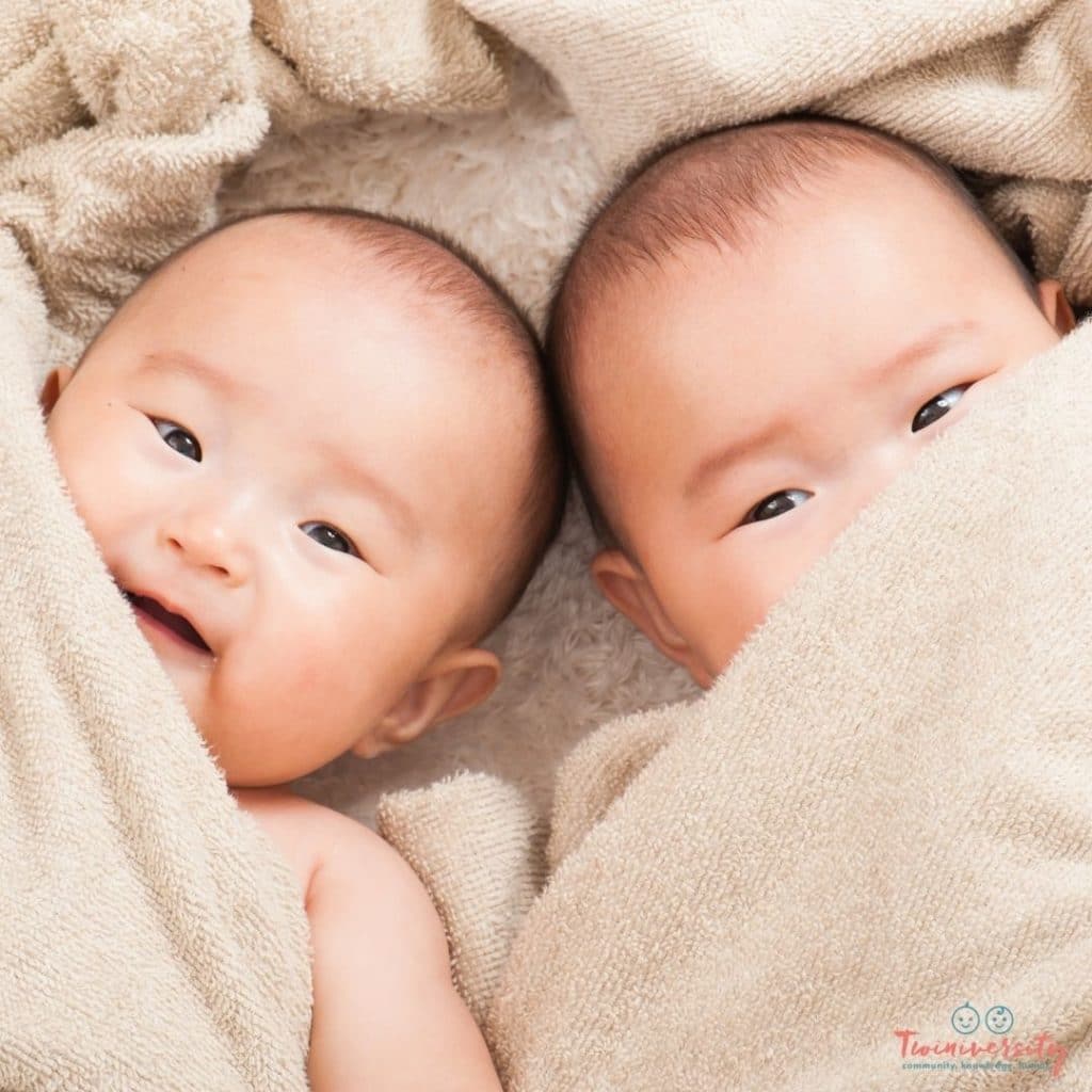 Identical twin babies lying head to head, on a fuzzy tan blanket. The baby on the right is peeking out from under the blanket and the baby on the left is smiling.