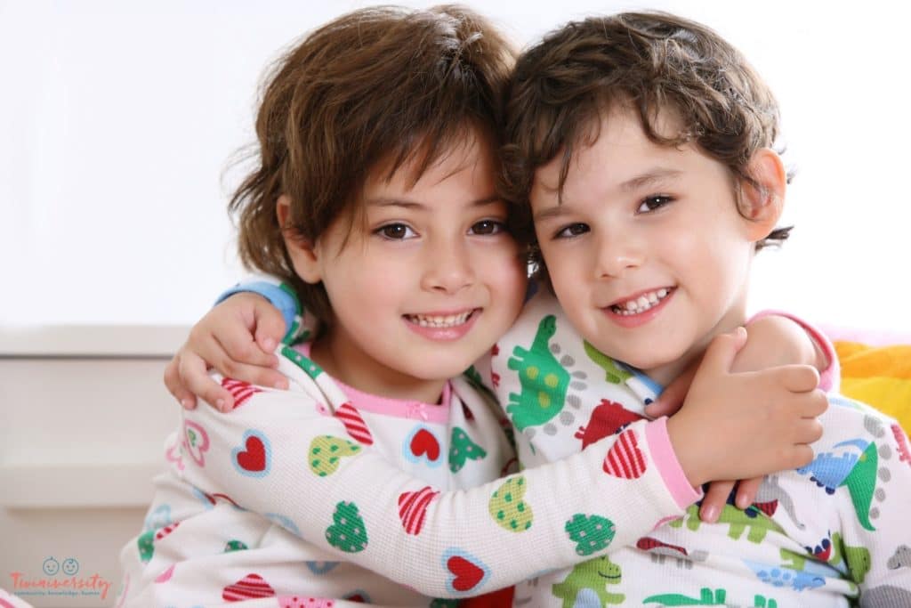 Girl/Boy twins side hugging each other. The girl is wearing heart pj's and the boy is wearing dinosaur pj's.