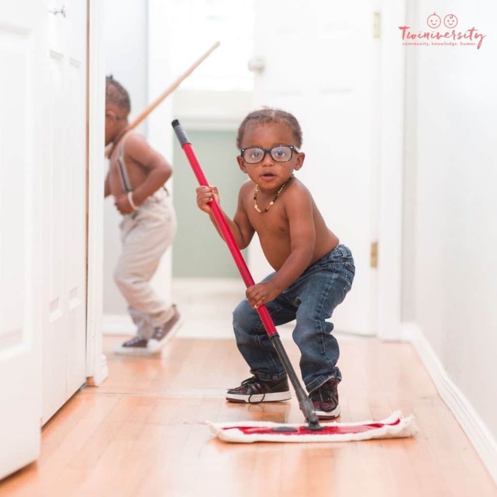 Twin boys are helping clean the hallway floor with big swiffer-sweepers. 