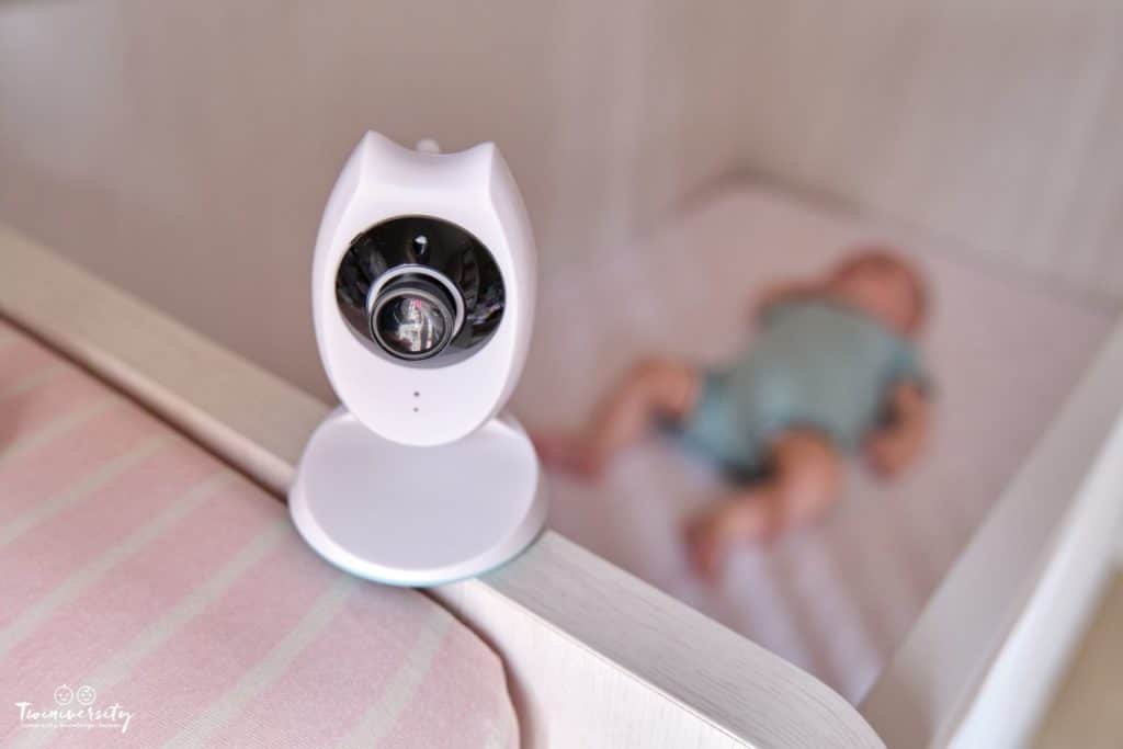 Baby monitoring camera is in the forefront of the image, resting on the side of the crib. Sleeping baby in a teal onesie is out of focus in the background