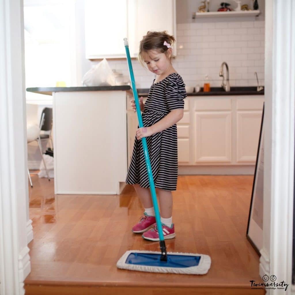 A young girl in a black and white striped dress with pink sneakers on is dry mopping the kitchen hardwood floor.