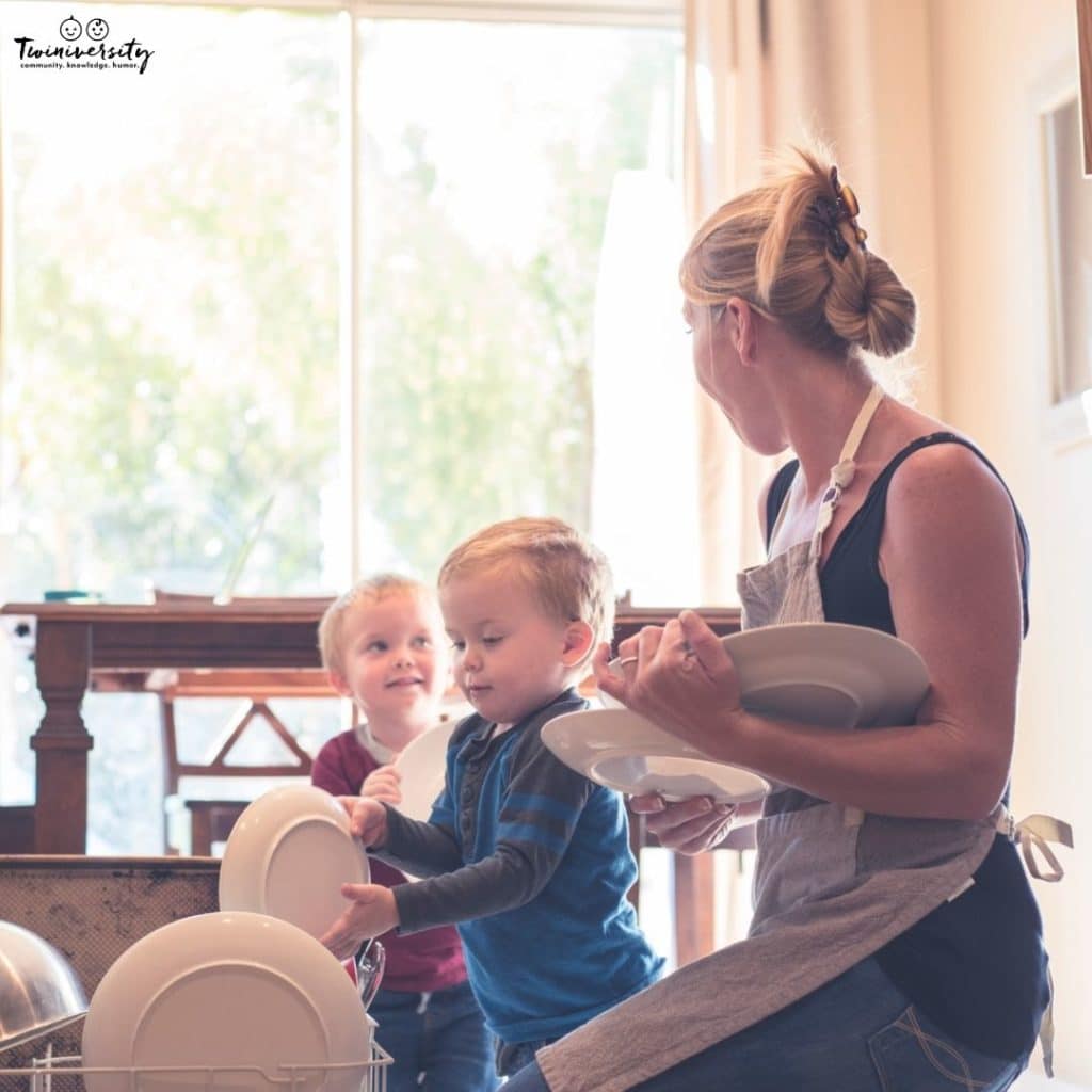 A mom with toddler boy twins is loading the dishwasher with plates and teaching her children responsibilities.