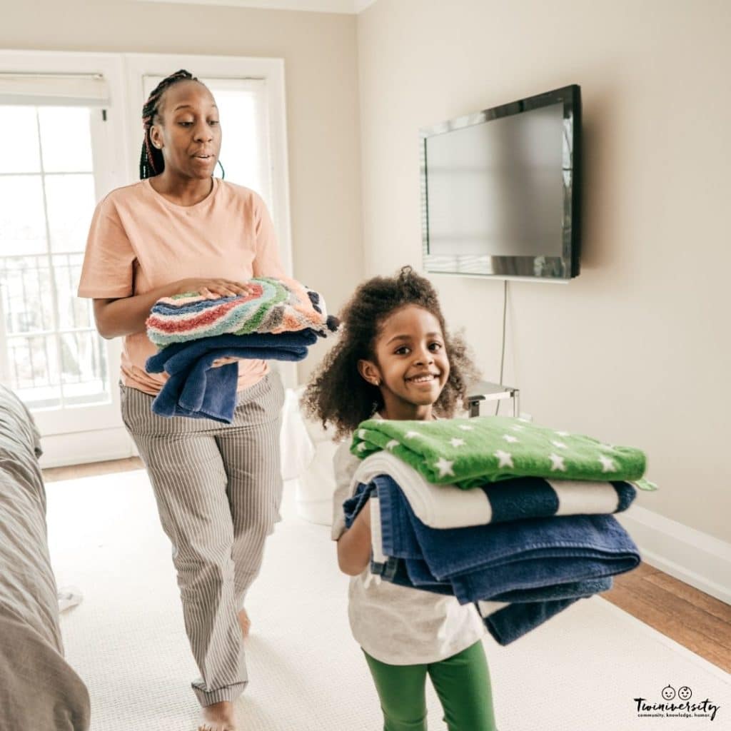 A mom and young daughter have been folding laundry and are now carrying them to put away.