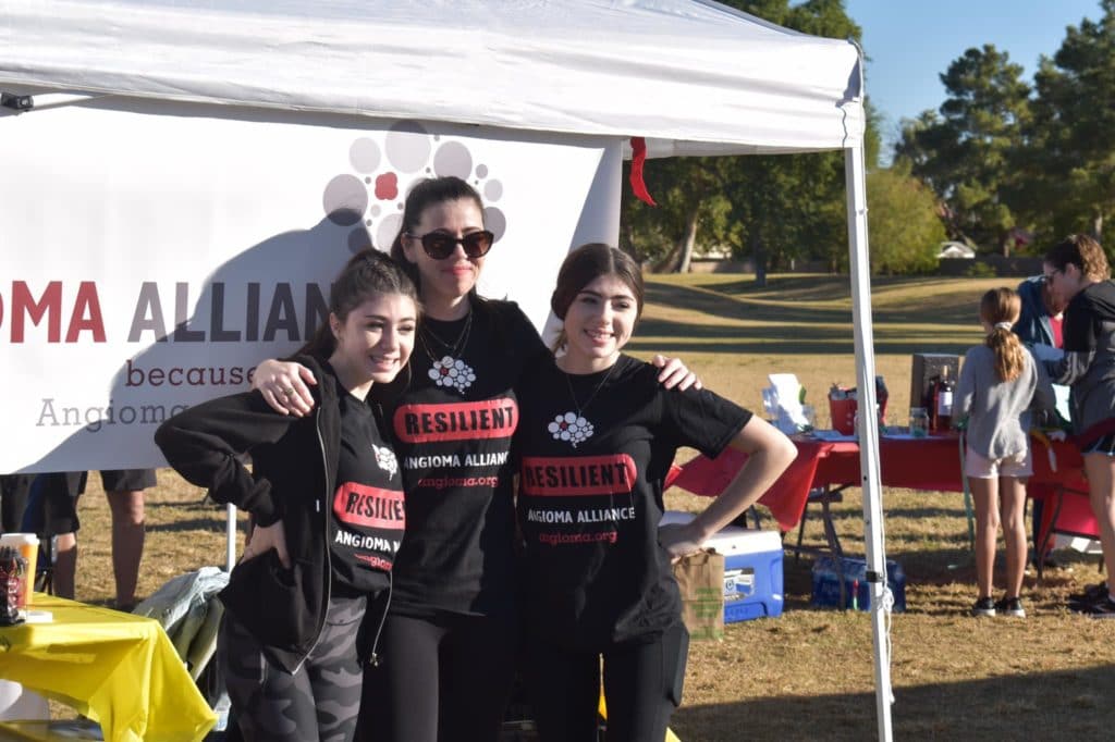 a woman standing with twin girls all in matching black shirts with the words "resilient" and "angioma alliance" on it standing outside in front of a banner.