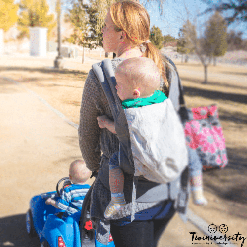 Mother managing twins solo on a walk to the park with one on push car and one in carrier
