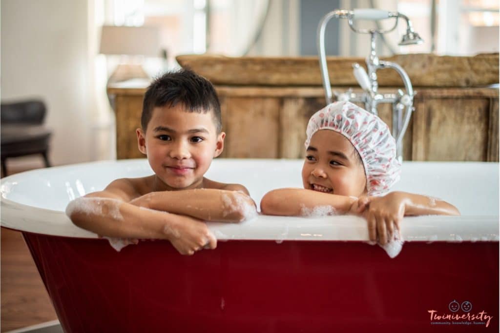 Young boy-girl siblings in a red bathtub together. Both are wet and covered in bubbles, with smiles on their faces.