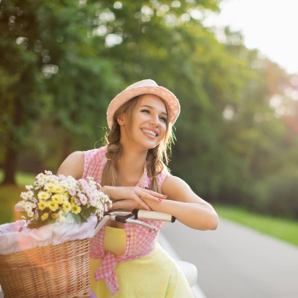 A woman is on a bicycle, leaning on the handlebars smiling. Her bike has a front basket that is carrying flowers.
