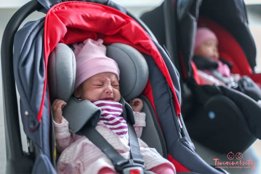 Twin baby girls wearing pink caps strapped into car seats. One baby is sleeping and one baby is upset.