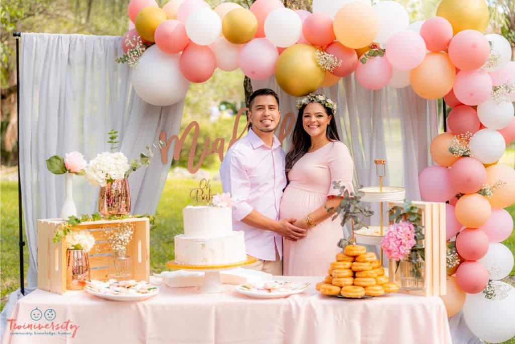 Latin man and pregnant woman standing behind the cake table at their baby shower. They are surrounded by a balloon arch, drapery and yummy treats.