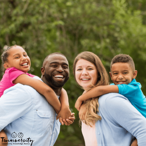 a family with one type of twins smiles at the camera outside as kids hang on parents' backs