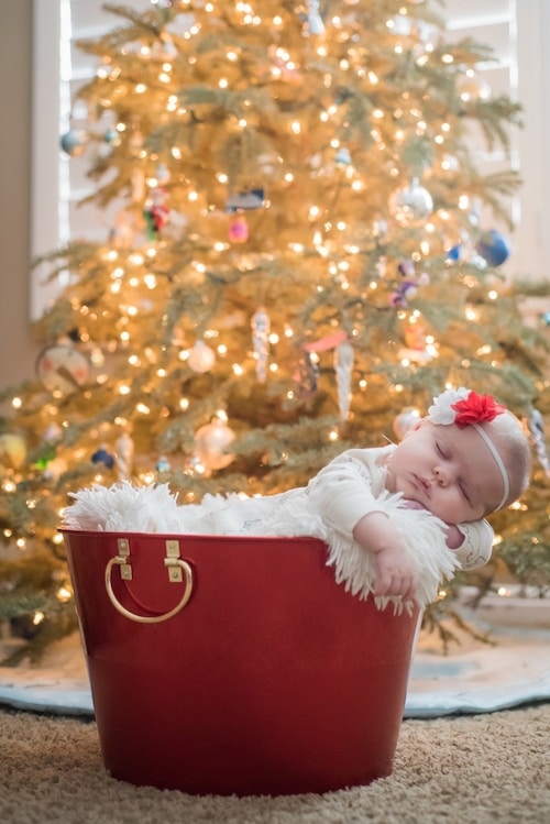 baby sleeping in a bucket set up in front of christmas tree 