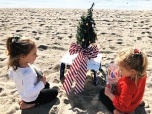 two young girls sitting on the beach with a tiny christmas tree new traditions