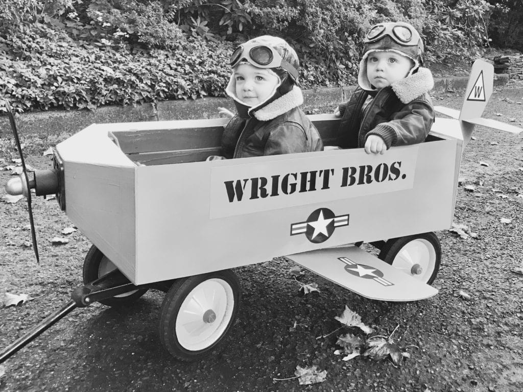 twin toddler boys dressed as the Wright brothers and sitting in a wagon decorate to look like an airplane