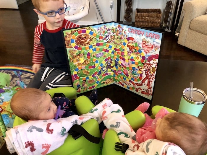 boy holding up candy land board to his infant twin sisters choose toys