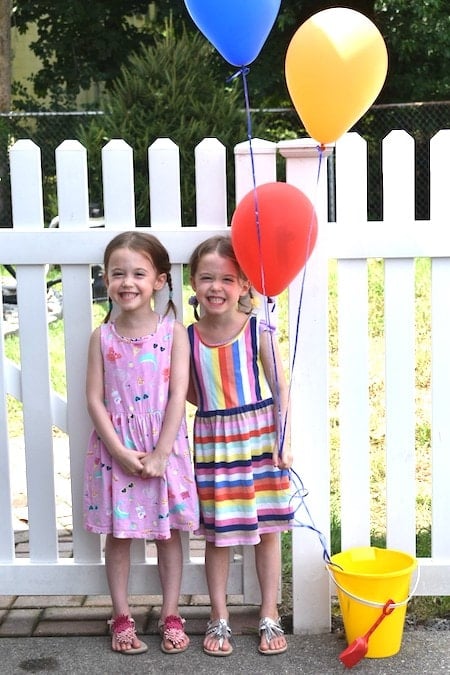 twin 5 year old girls standing in front of white picket fence with balloons embrace their individuality