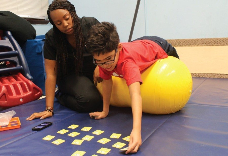 occupational therapist with boy laying on an exercise ball and playing a card game on a mat 