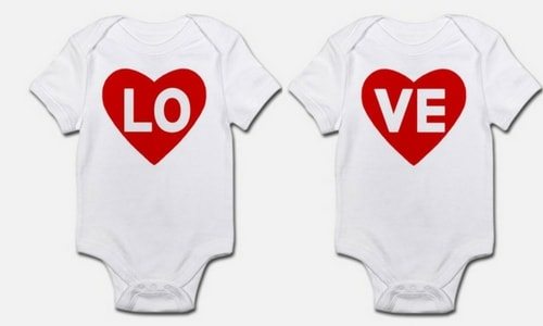 Two white baby onesies side by side, the left one with a red heart and white letters "L" and "O", the right with a matching red heart and white letters "V" and "E." used for a Valentine's day baby announcement.