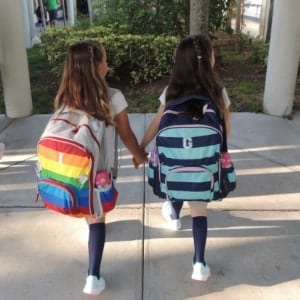 twin girls holding hands and walking on a sidewalk with backpacks on