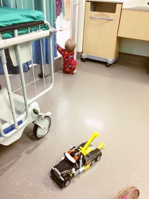 1 year old boy at the glass door of a hospital room in pajamas ear tubes