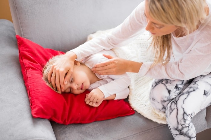 girl lying on couch sick with mom touching her forehead the flu