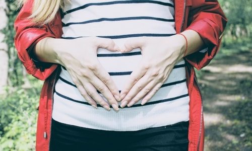 Pregnant woman, pictured from chest to hips, with red raincoat, black and white striped sweater, and black pants, holding hands in shape of a heart over belly