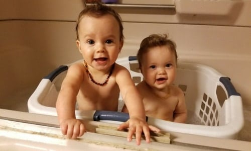Twin toddlers sitting in a white laundry basket placed in the bathtub.