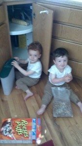 two boys sitting on a floor with a bag of cereal in front of an open cabinet