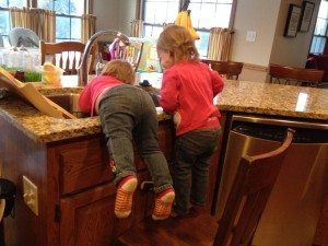 twin toddlers climbing onto a sink with a chair in a kitchen
