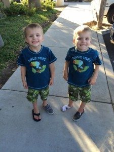 young twin boys in matching outfits, standing outside on a sidewalk next to each other, smiling at the camera