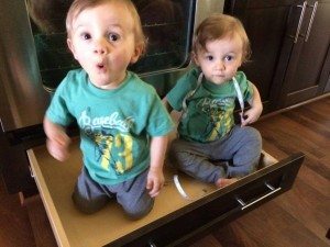 twin toddler boys dressed alike creating mischief by sitting in a kitchen drawer