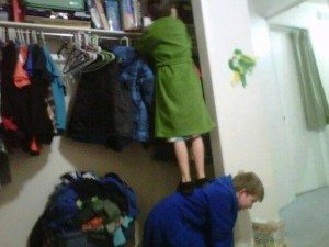 one kid in a bathrobe climbing on the other kids back to reach into a closet shelf
